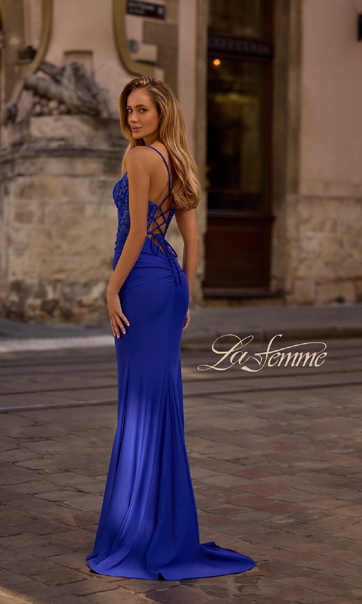 Woman in a fitted royal blue Lace-Up Open-Back Long Prom Dress (La Femme 32217), standing on a cobblestone street in front of an old building;